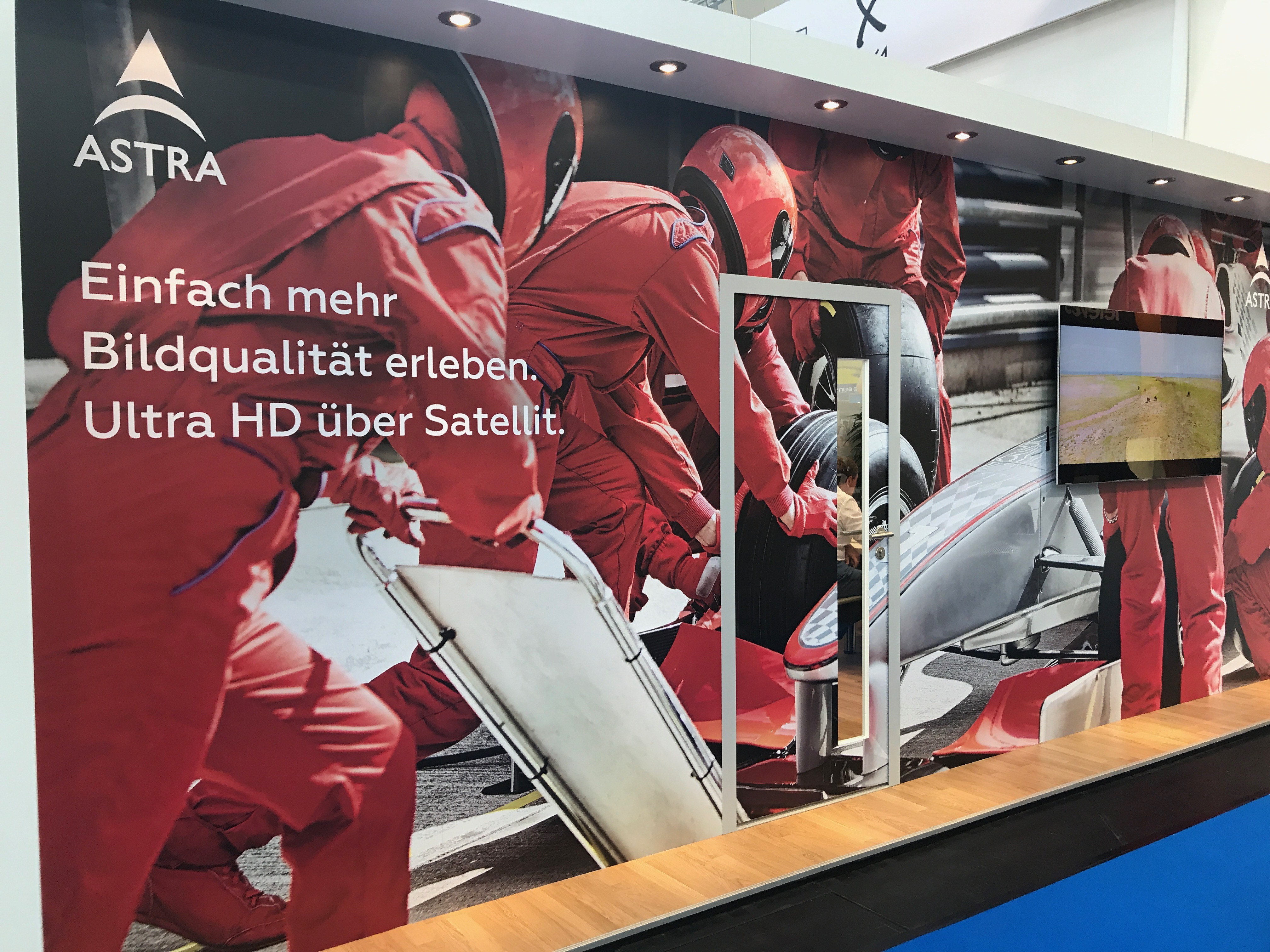 Messestand von ASTRA mit Werbeslogan für Ultra HD über Satellit und einem Formel-1-Boxenstopp-Motiv im Hintergrund